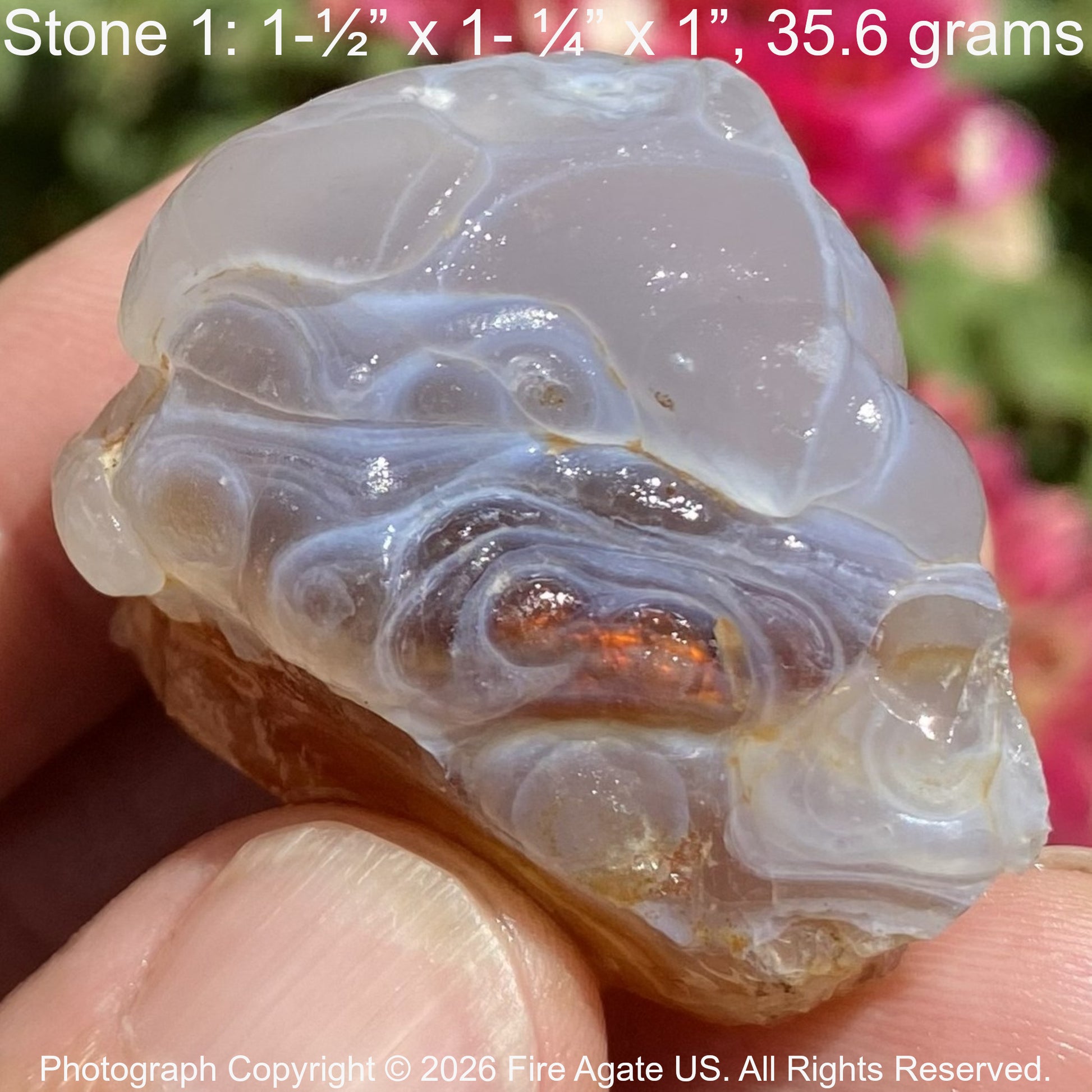 A hand holds a fire agate stone specimen with swirling translucent bands in white, gray, and pale blue, showing a small amber orange inclusions near the center. Text at the top reads 'Stone 1: 1-1/2 x 1-1/4 x 1", 35.6 grams'