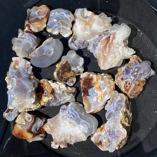 A group of raw fire agate stone slices and chunks with banded patterns in white, gray, brown, and blue, arranged on a black plate.