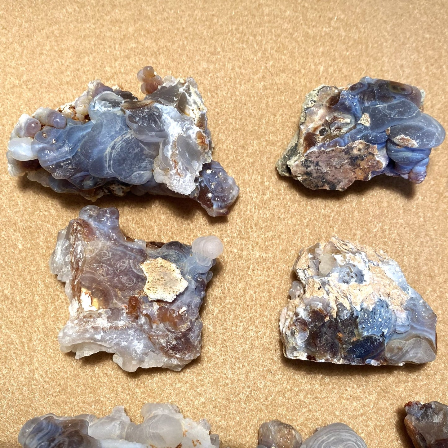 Four rough fire agate stones arranged on a tan textured background. The stones show layered translucent bands in gray, blue, white, and brown with some metallic-looking crystalline areas.