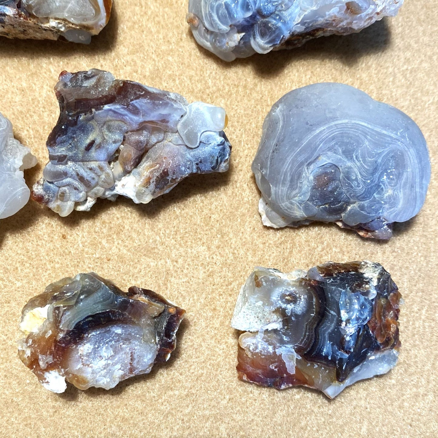 Four raw fire agate stone pieces with natural banding and rough, unpolished edges arranged on a tan textured background.