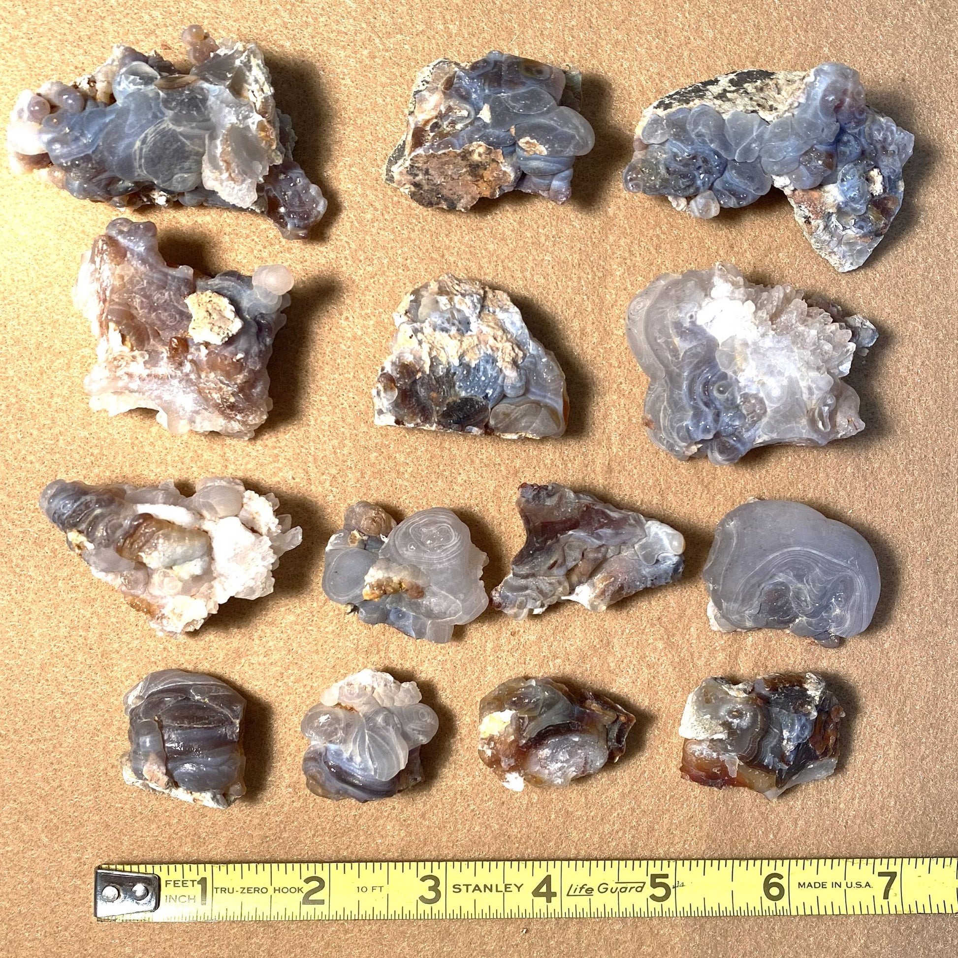 Fourteen pieces of Slaughter Mountain fire rough agate arranged in a grid on a tan background, with a yellow Stanley tape measure along the bottom edge for scale.