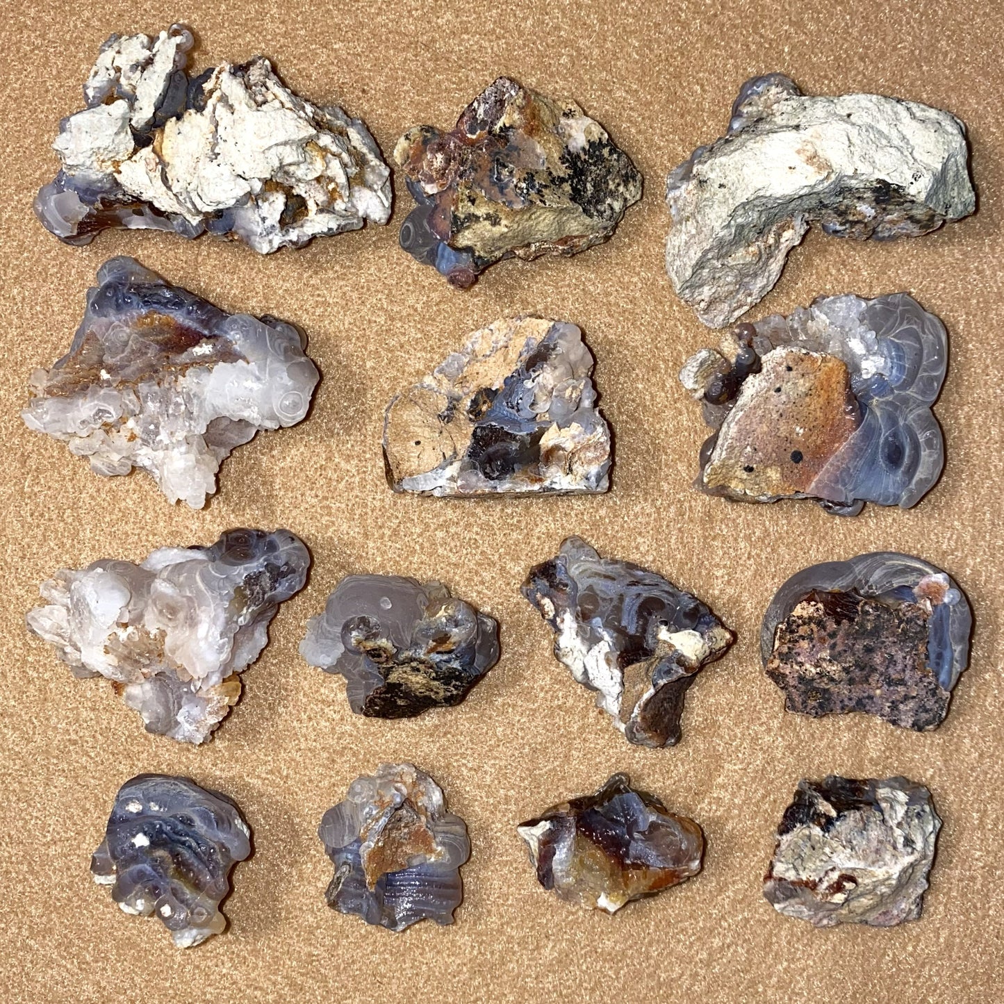 A set of fourteen raw fire agate and quartz crystal specimens arranged in a 3x4 grid on a textured tan background.