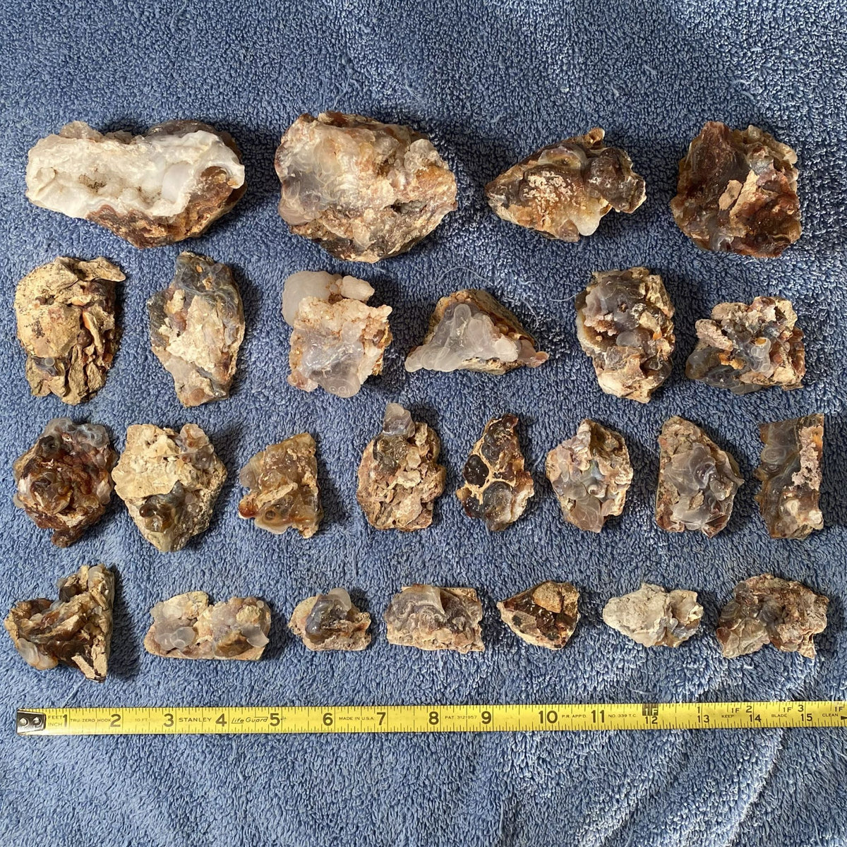 A group of rough raw fire agate with quartz crystal clusters arranged in rows on a blue textured blanket, with a yellow measuring tape at the bottom indicating size.
