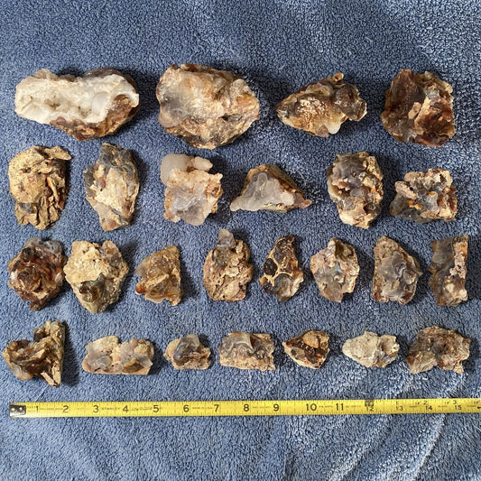 A group of rough raw fire agate with quartz crystal clusters arranged in rows on a blue textured blanket, with a yellow measuring tape at the bottom indicating size.