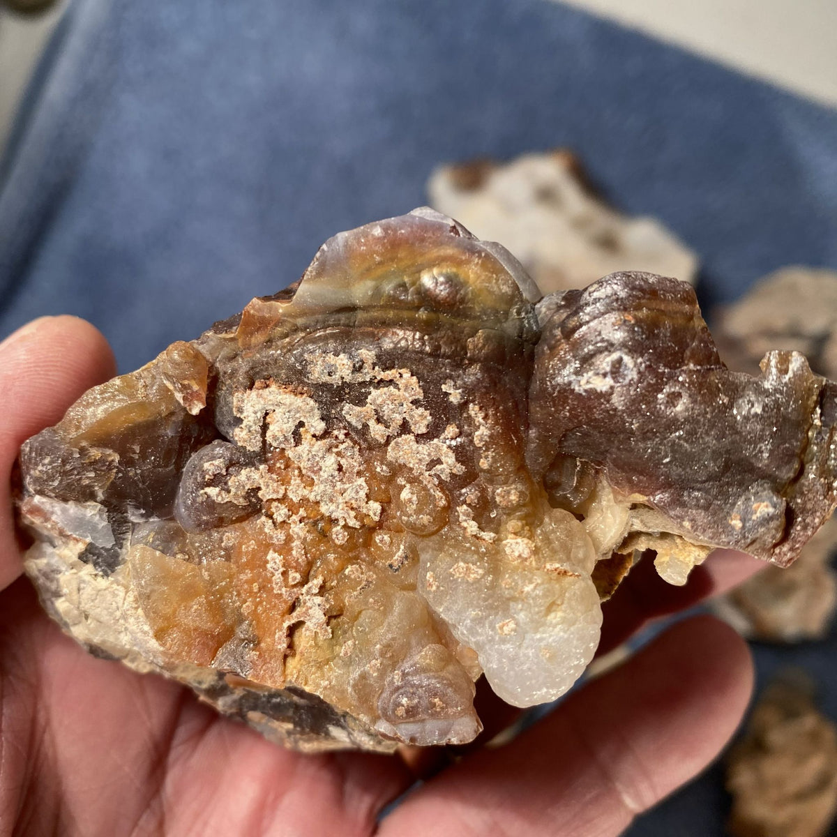 A hand holding a rough natural fire agate stone with layered translucent crystals and metallic-looking brown and amber tones, against a blue fabric background.
