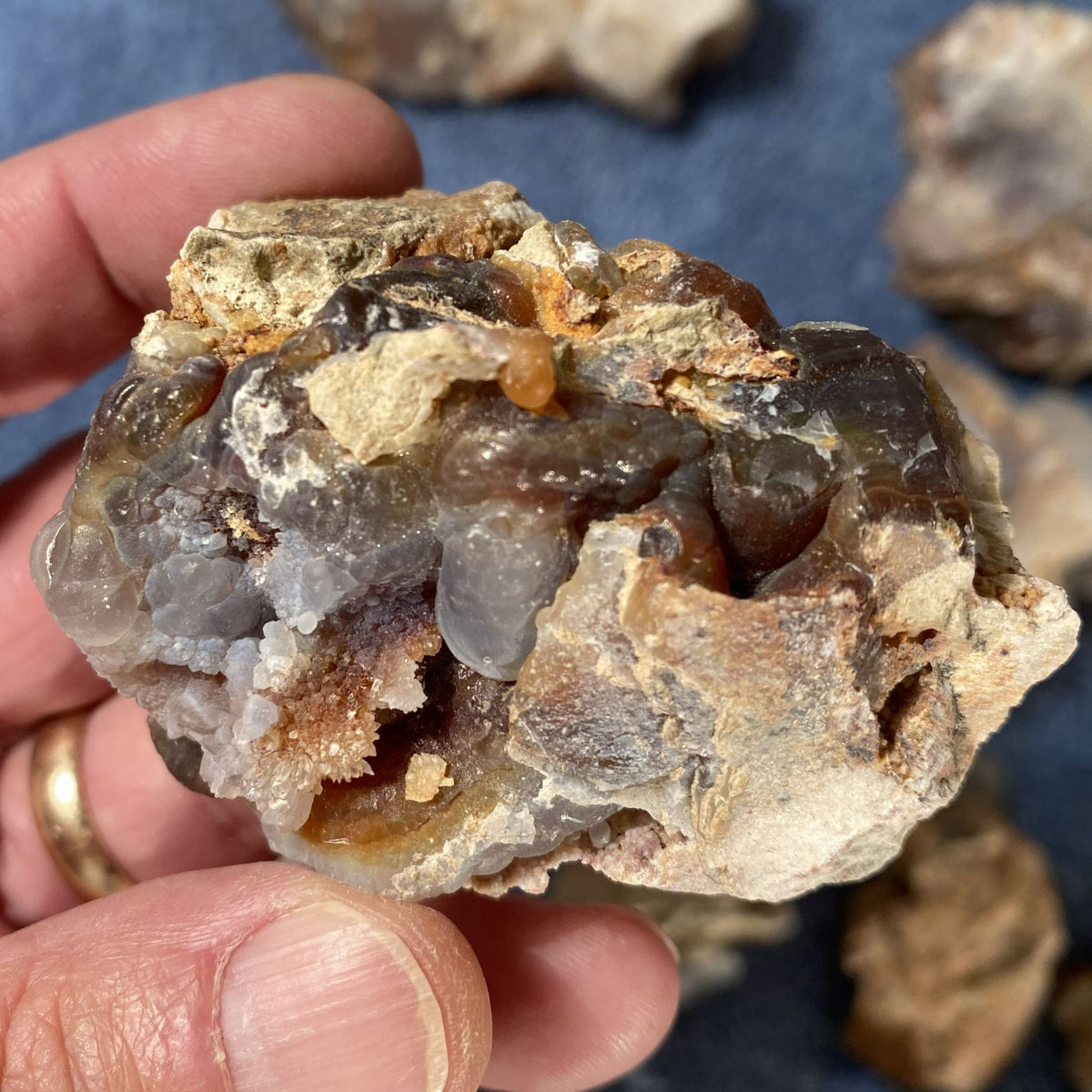 A hand holding a piece of fire agate rough with translucent gray and brown crystals on a textured beige matrix, with other similar specimens blurred in the background.