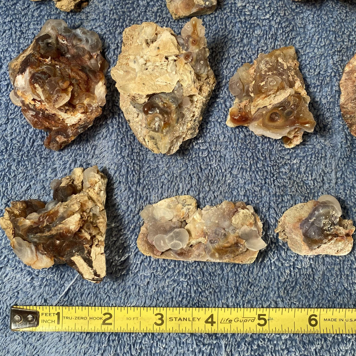 Several raw fire agate stone specimens with visible internal cavities and crystalline formations are arranged on a blue textured towel, with a yellow Stanley tape measure along the bottom for scale.