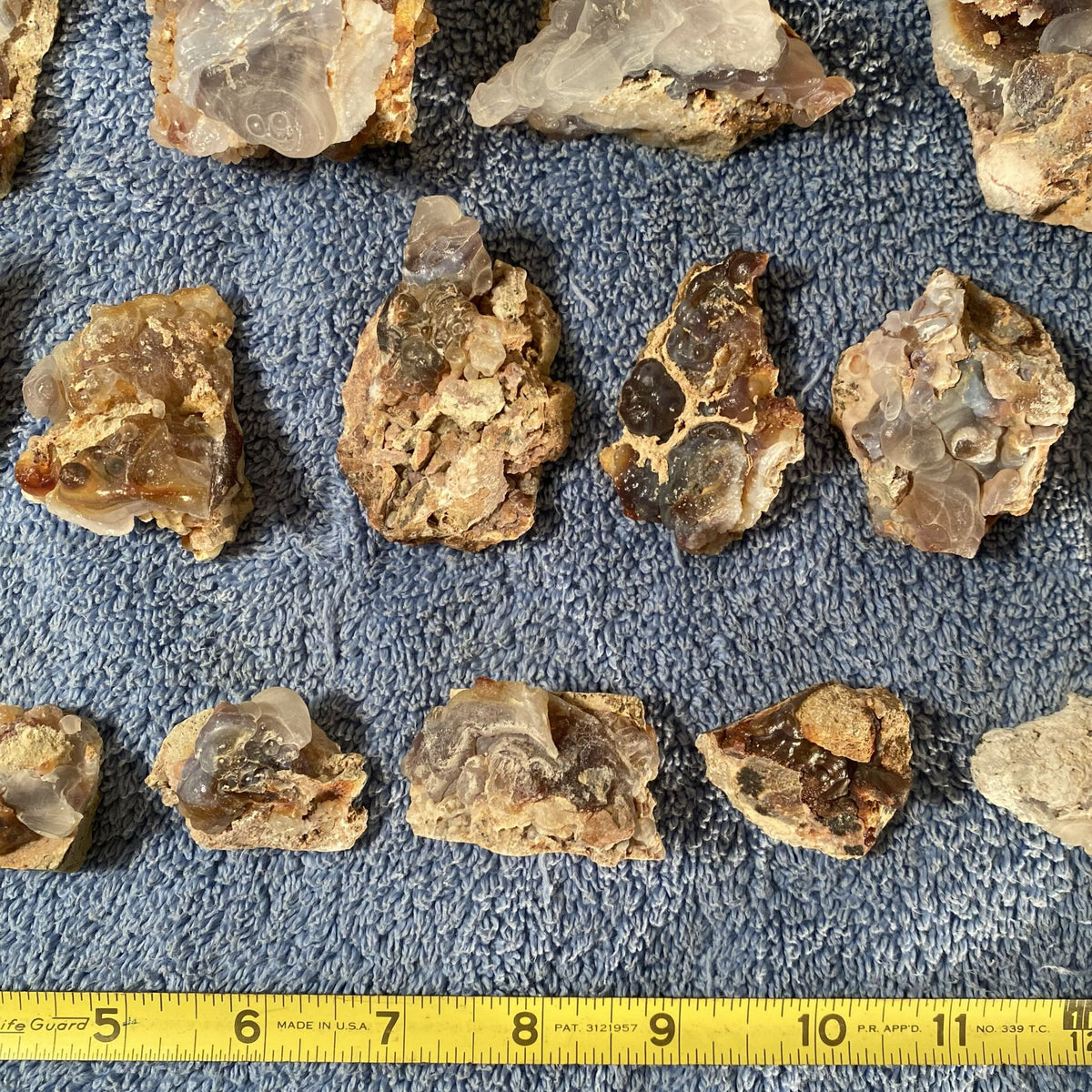 Several rough translucent fire agate crystal/mineral specimens arranged on a blue textured surface, with a yellow tape measure along the bottom edge for scale.
