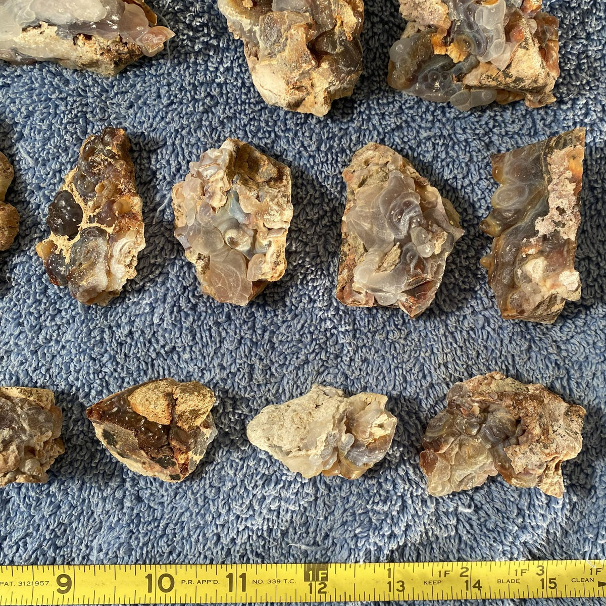 Several rough fire agate stone chunks with layered translucent bands are arranged on a blue textured towel, with a yellow tape measure visible along the bottom edge for scale.