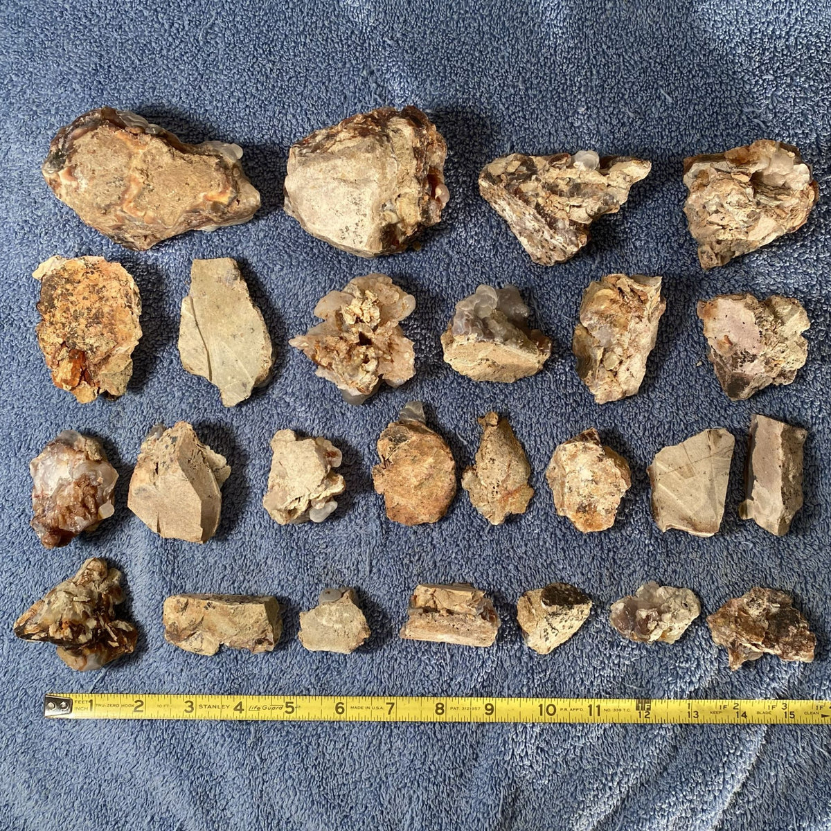 A collection of assorted natural fire agate stones arranged in rows on a textured blue fabric surface, with a yellow tape measure placed along the bottom edge for scale.